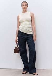 Kvinna i ärmlös beige topp, vida mörkjeans, svarta sandaler, håller en liten brun väska, står mot en enfärgad vit vägg.