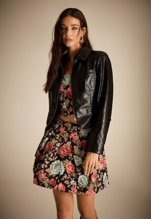 Femme portant une veste en cuir noire sur un crop top à motif floral et une jupe assortie, debout devant un fond beige uni.