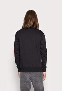 Sudadera negra con puños y dobladillo acanalados, cuenta con un bolsillo adicional y un detalle rojo en una manga, hecha de una tela suave y texturizada.