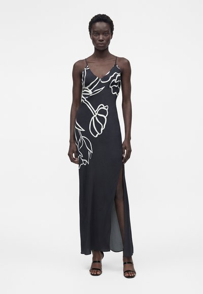 Γυναίκα που φοράει μαύρο μάξι φόρεμα χωρίς μανίκια με λευκές floral περιγράμματα και σκίσιμο ψηλά στο μπούτι, συνδυασμένο με μαύρα σανδάλια με λουράκια.