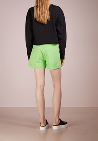 Shorts verts en tissu lisse, dotés d'une coupe classique, de poches arrière et d'une ceinture. Associés à un sweat-shirt noir et des chaussures noires à enfiler.