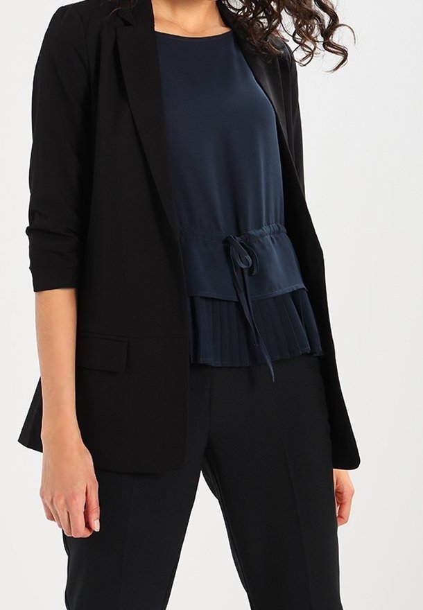 Blazer noir sur mesure porté sur un top peplum bleu foncé avec un cordon de serrage. Le pantalon est noir, complétant la tenue professionnelle.