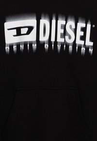 Fekete kapucnis pulóver, fehér "DIESEL" logóval, amelynek színátmenetes hatása van, alatta a "DENIM DIVISION" felirattal. Elöl zseb található.