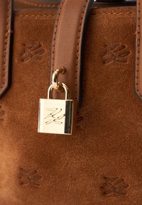 Καφέ σουέτ τσάντα χειρός με χρυσό λουκέτο και ανάγλυφο λογότυπο. Ο σχεδιασμός περιλαμβάνει καφέ δερμάτινες λεπτομέρειες και λεία υφή.