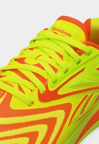 Κοντινό πλάνο νεον κίτρινου και πορτοκαλί παπουτσιού Reebok με κορδόνια και λογότυπο στη γλώσσα, που δείχνει λεπτομερή υφή και σχέδιο.