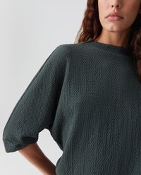 Vrouw met golvend bruin haar draagt een getextureerde donkergroene top met vleermuismouwen, staande tegen een effen lichte achtergrond.
