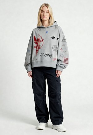 Donna bionda che indossa una felpa con cappuccio grigia con cupido rosso, occhi e testo "Je T'aime", abbinata a pantaloni cargo neri e sneakers bianche, in piedi.