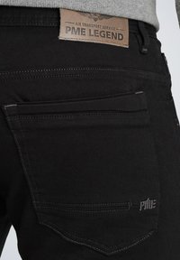 Čierne džínsy z denimu s hnedou koženou nášivkou s nápisom "PME LEGEND." Medzi vlastnosti patrí zadné vrecká s nenápadným prešívaním a štruktúrovaný povrch.