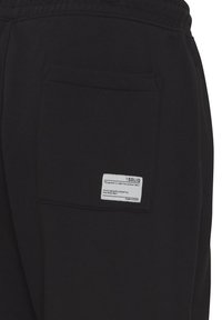 Zwarte joggingbroek met een achterzak en een wit etiket. De stof lijkt zacht en duurzaam, met een gladde textuur.