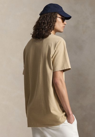 Άτομο με σγουρά μαλλιά που φοράει μπεζ μπλούζα, σκούφο σκούρου μπλε, γυαλιά ηλίου και λευκό παντελόνι, στέκεται με το ένα χέρι στην τσέπη, στραμμένο πλαγίως.