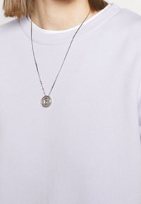 Sweatshirt lilas avec un col rond, en tissu texturé. Un collier argenté avec un pendentif circulaire repose sur le col.