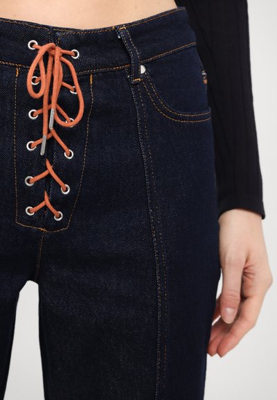 Los vaqueros de denim oscuro cuentan con un frente con cordones en color naranja, ojales plateados y costuras en contraste. Los bolsillos son visibles a cada lado.
