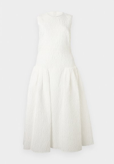 Bela maxi obleka brez rokavov s teksturirano tkanino, oprijetim pasom in razširjeno, naborki oblikovano krilo na enobarvnem ozadju.