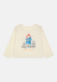 Bela majica s dolgimi rokavi iz bombaža, ki prikazuje risanega medvedka v modrem plašču in rdeči kapi, obkroženega s hrano ter besedilom "Paddington".