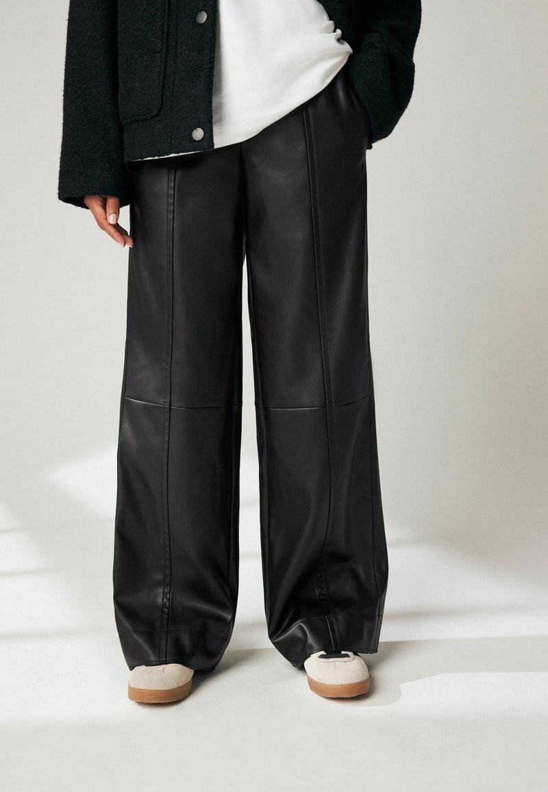Des pantalons larges en simili cuir noir, dotés d'une texture lisse, de coutures marquées et d'une coupe décontractée, associés à des chaussures de couleur claire.