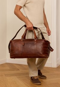 Braun kožnati putni torba s dvostrukim ručkama, remenom za rame i prednjim džepom na zatvarač. Tekstura s vidljivim detaljima šivanja.