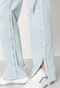 Jeans de ganga azul claro com detalhes de desgaste nos joelhos e aberturas laterais. Combinados com sapatilhas brancas com detalhes azuis.