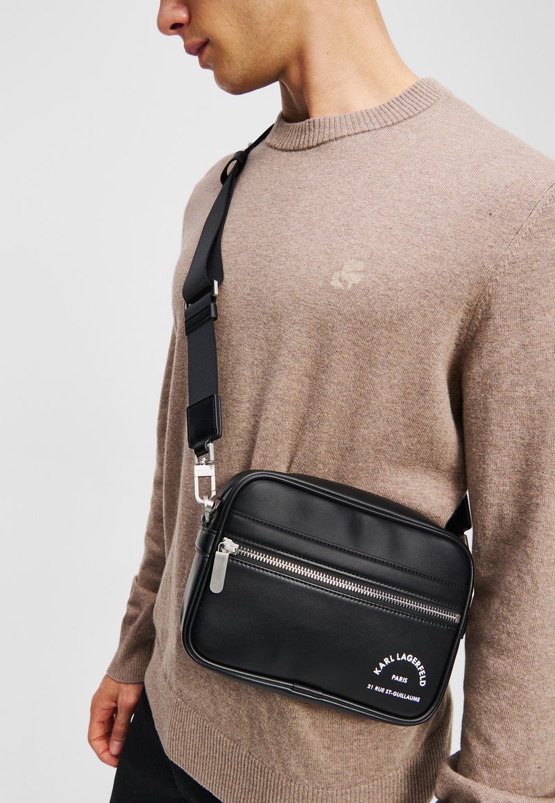 Black leather crossbody bag with silver hardware, zippered compartment, and white logo text. Features an adjustable strap and rectangular shape.