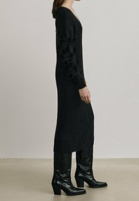 Black knitted dress with long sleeves, V-neck, and fringe detailing on sleeves. Paired with knee-high black heeled boots.
