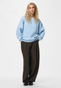 Femme aux longs cheveux blonds bouclés portant un sweat-shirt bleu clair surdimensionné, un pantalon ample marron foncé et des chaussures noires, debout les mains dans les poches.