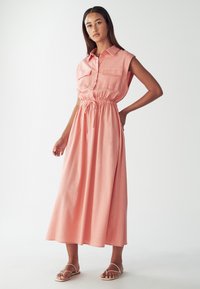 Bežė su rankovėmis, apykaklės suknelė iš minkšto rožinio audinio, su suveržta juosena ir dviem priekiniams kišenėms, tekanti iki kelių.