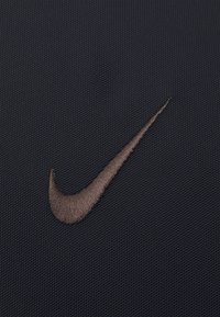 Nike-logotyp på mörk tyg, med en broderad swoosh i metalliskt brons. Texturerad yta med ett fint nätmönster.