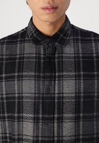 Muž v černobílém kostkovaném košili s knoflíky, límec zapnutý, zobrazený od brady po hruď.