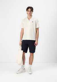 Camicia a maniche corte di colore crema con una texture sottile e un piccolo logo giallo, indossata con pantaloni corti scuri e sneakers bianche.