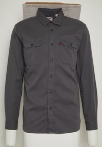 Mörkgrå långärmad skjorta i bomull. Har knapp-up framtill, två bröstfickor och en liten röd etikett på vänstra fickan.