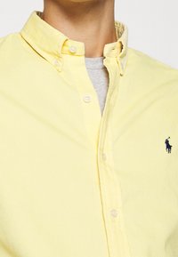 Žlutá košile na knoflíky s límečkem, s modrým vyšitým logem na levé straně hrudi a s texturovaným detailem látky.