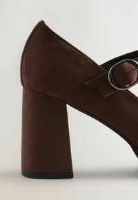 Pantof  cu toc înalt din suede maro, având un toc gros și o curea cu cataramă pe lateral. Designul include un vârf ascuțit și o textură netedă.
