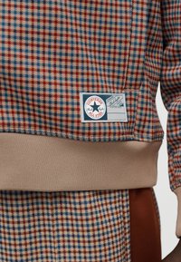 Φούτερ με καρό σχέδιο σε  κόκκινο, μπλε και κρεμ, με μπεζ ριμπ τελείωμα. Περιλαμβάνει ετικέτα με το λογότυπο «Converse Chuck Taylor».