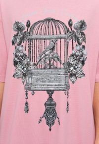 Ροζ μπλουζάκι με λεπτομερή εκτύπωση σε μαύρο και άσπρο ενός κλουβιού πουλιών, ενός πουλιού και λουλουδιών με διακοσμητικά στοιχεία και περίπλοκα μοτίβα.