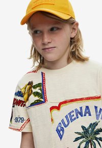 Beige t-shirt med färgglada broderade mönster, som visar palmer och text. Kombinerad med en ljusgul keps.