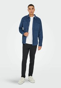 Blå kordfløjl skjorte med knap-op design, to brystlommer, båret over en hvid t-shirt, parret med sorte bukser og hvide sneakers.