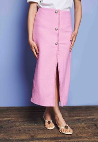 Γυναίκα με ροζ φούστα ψηλής μέσης και κουμπιά, που έχει σκίσιμο μπροστά και λευκά σανδάλια, στέκεται σε ξύλινο δάπεδο μπροστά σε μπλε τοίχο.