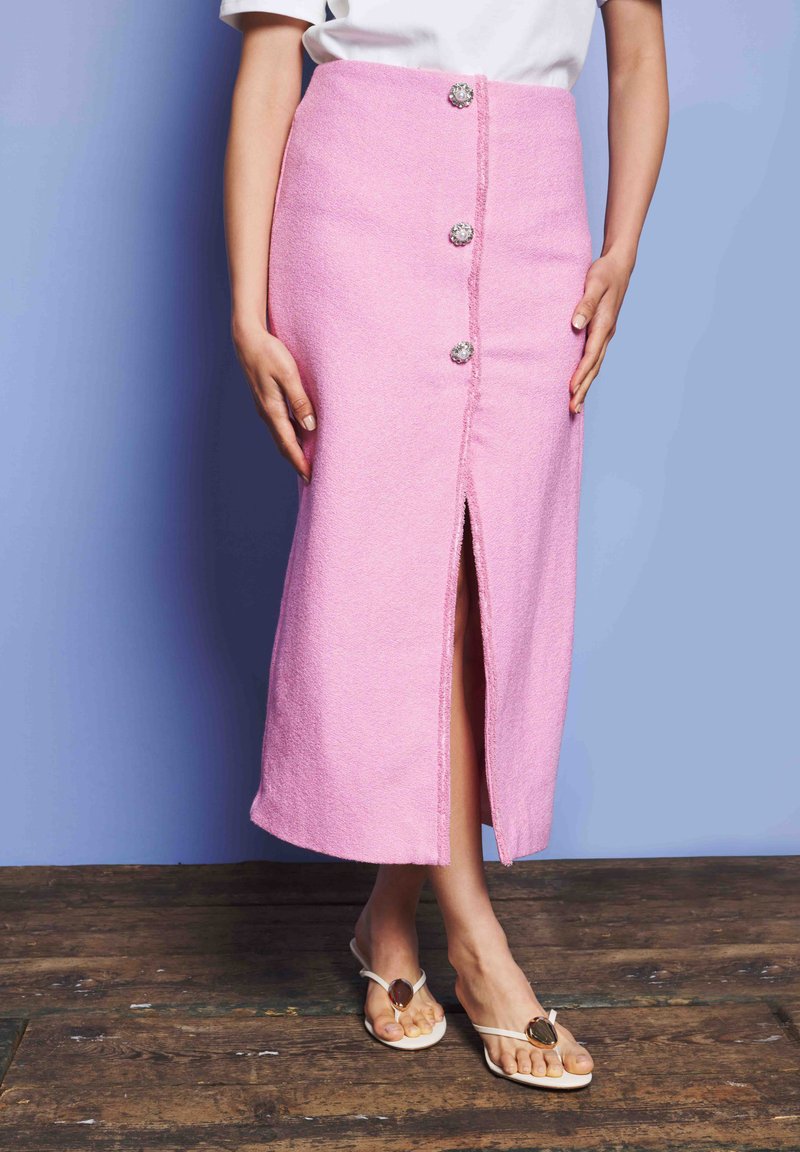 Γυναίκα με ροζ φούστα ψηλής μέσης και κουμπιά, που έχει σκίσιμο μπροστά και λευκά σανδάλια, στέκεται σε ξύλινο δάπεδο μπροστά σε μπλε τοίχο.