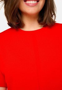 Haut en tricot rouge avec un col rond, des manches courtes et une texture lisse. Détail de couture notable au centre.