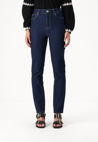 Ψηλόμεσα σκούρα μπλε τζιν με αντίθεση ραφής, πέντε τσέπες και κομψό, κομμένο σχέδιο ποδιού, συνδυασμένα με μαύρα σανδάλια που διαθέτουν μεταλλικούς προσαρμογείς.