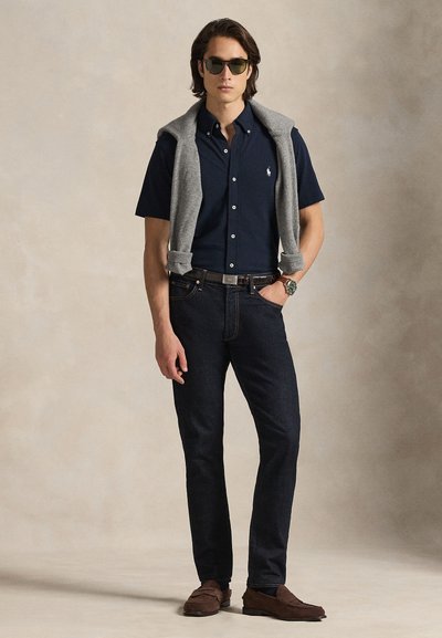 Μπλε πουκάμισο με κουμπιά, γκρι πουλόβερ ριγμένο στους ώμους, σκο ύρο τζιν, καφέ μοκασίνια και γυαλιά ηλίου, με ασημένιο ρολόι στον καρπό.