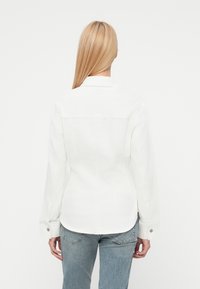 Valge teksajope pikkade varrukatega, terava krae, vormitud lõike ja metallist tõmmiknuppudega. Tagantvaade näitab siledat tekstuuri ja õmblusdetailide.