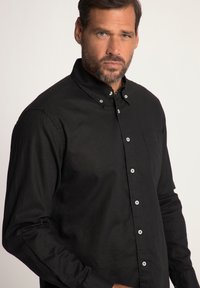 Camisa negra de botones con collar clásico, mangas largas, bolsillo en el pecho y botones blancos contrastantes; la tela parece texturizada.