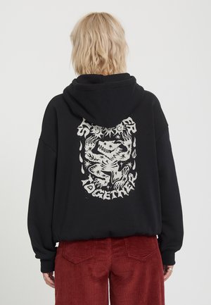 Person mit kurzem blondem Haar trägt einen schwarzen Hoodie mit weißer Grafik und Text auf dem Rücken, kombiniert mit roten Cordhosen.