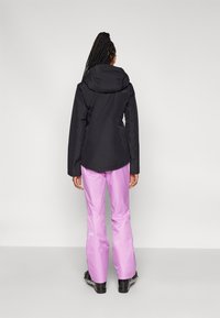 Zwarte waterdichte jas met een capuchon, gecombineerd met lichtpaarse waterdichte broek. De outfit heeft een ontspannen pasvorm en is geschikt voor buitenactiviteiten.
