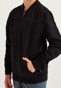 Czarna kurtka bomberka z zamkiem z przodu, ściągaczami przy mankietach oraz kieszeniami po bokach; ma teksturowany materiał i delikatne logo na piersi.