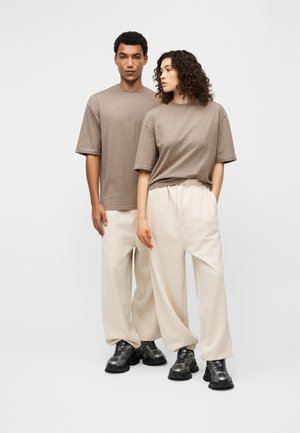 Δύο μοντέλα φορούν ταιριαστά μπεζ-γκρι t-shirts, μπεζ πλαδιές παντελόνες και μαύρα χοντρά αθλητικά παπούτσια, στέκονται μπροστά σε απλό λευκό φόντο.