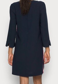 Marineblaues Kleid mit Dreiviertelärmeln, seitlichen Schlitzen und kontrastierenden roten Streifen entlang der Seitennähte. Glatte Stoffqualität, gerafftes Rückendetail.