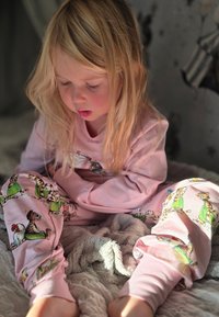 Light pink pajama set featuring whimsical animal illustrations in various colors. Material appears soft; child sits on a textured gray blanket.