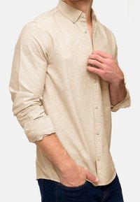 Chemise beige à manches longues avec col boutonné, une poche poitrine unique et des poignets retroussés, fabriquée en tissu texturé.