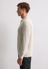 Hellfarbener, strukturierter Strickpullover mit Polokragen, langen Ärmeln und geripptem Saum, kombiniert mit dunklen Hosen, seitlich betrachtet.